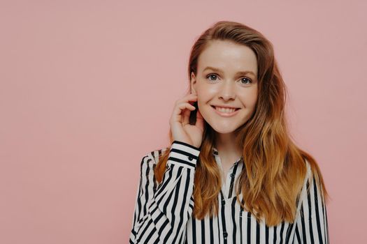 Positive young woman in stripy black and white blouse with wavy ginger hair touching face, smiling at camera and posing over light pink studio background