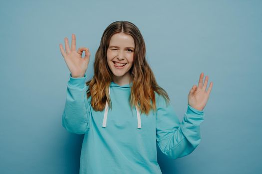 Excited teenage girl with wavy ombre hair showing okay sign closing one eye and sticking tongue out while posing in casual wear on studio background. Body language concept