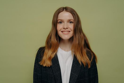Portrait of smiling young woman with wavy medium length ombre hair wearing dark formal jacket and white top posing isolated over green studio background