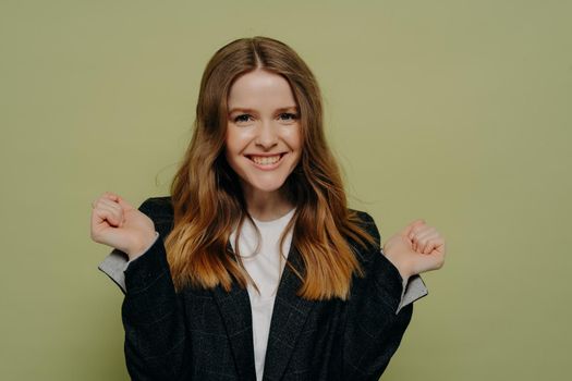 Happy young female with wavy brown hair holding hands up demonstrating excitement wearing dark formal jacket and white top, posing against light studio background. Human emotions concept