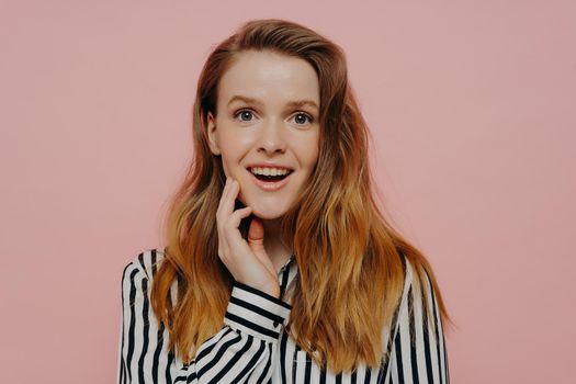 Amused young woman in striped black and white blouse with wavy ginger medium length hair touching face, expressing amazement while posing over light pink studio wall. Human emotions concept