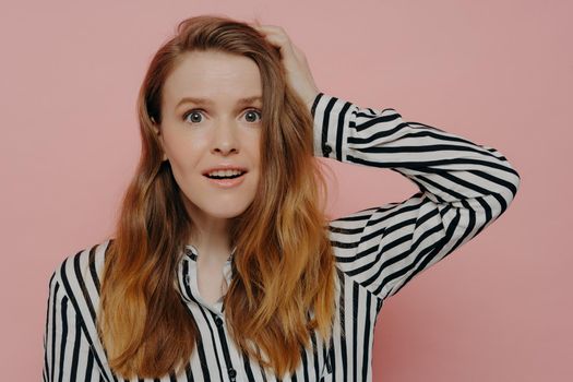 Attractive young female in stripy black and white blouse touching head with one hand in astonishment expressing surprise while posing against pink studio wall. Human emotions concept