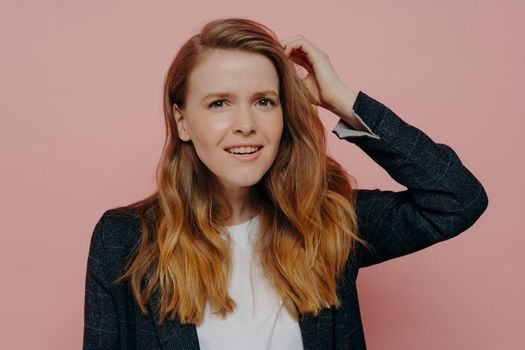Attractive pensive young woman with wavy ginger hair in formal dark jacket and white top thinking while touching head with confusion standing against pink studio background