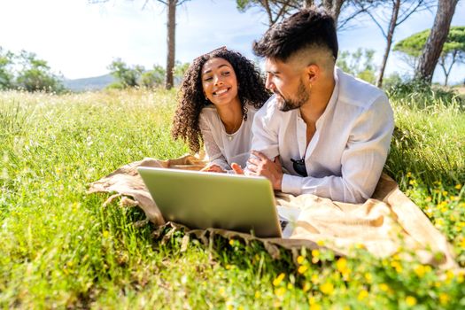 Happy couple in love using laptop in nature lying on a grass field sharing vacations on social network. Two millennial students working at pc in nature. New internet mobile connection technology