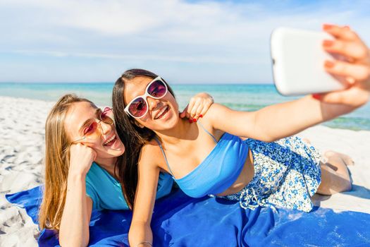 Two cute girls lying on white sand taking a self-portrait with cell wearing blue casual summer clothes and funny sunglasses on vacation at tropical beach resort with blue ocean sea on background