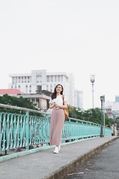 Full length of asian girl walking on a bridge with city view with a book in her hand