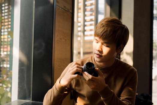 Asian Male professional photographer dressed in casual outfit making setting on display choosing filters on vintage camera and watching photos while resting in coffee shop interior near window