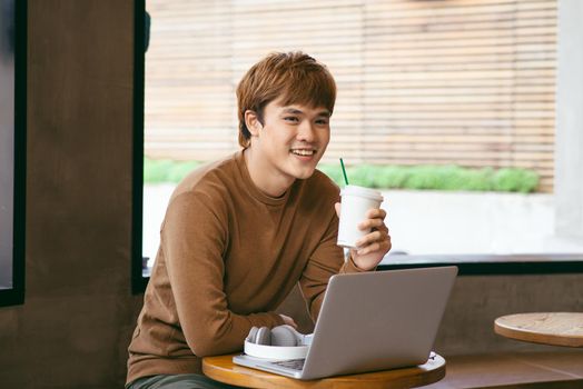 Handsome asian man using laptop sitting on wooden table in coffee shop with a cup of coffee