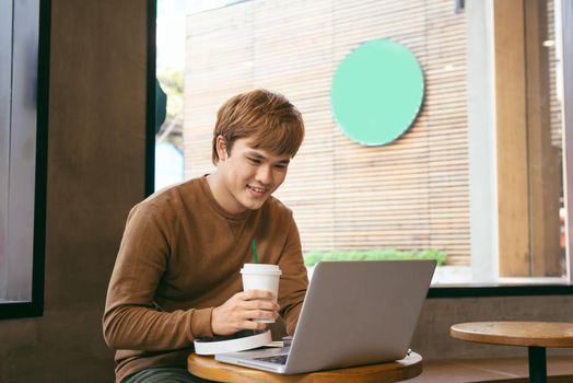 Smiling handsome worker in glasses drinking coffee while watching video on his laptop at office