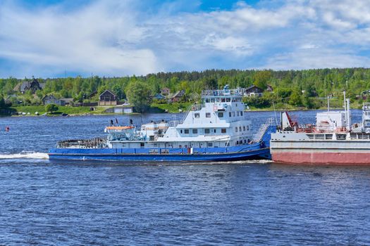 View of a tugboat pushing a heavy barge down the river along the shore. River transportation of goods