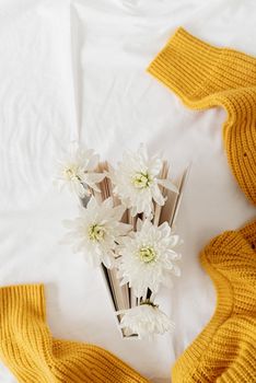 Spring reading concept. Top view of a book with white chrysanthemum flowers on white background with yellow sweater. Flat lay