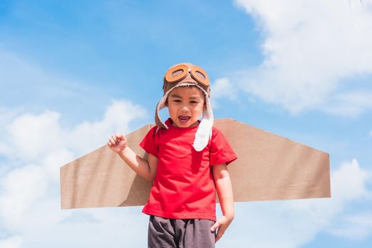 Happy Asian funny child or kid little boy smile wear pilot hat and goggles play toy cardboard airplane wing flying raises hand up against summer blue sky cloud background, Startup freedom concept