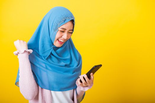 Asian Muslim Arab, Portrait of happy beautiful young woman Islam religious wear veil hijab funny smile she raise hand glad celebrating winning with smart mobile phone isolated on yellow background