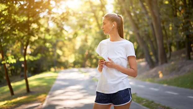Woman putting on earphones to listen music before jogging summer park Caucasian female preparing to run sunny morning Runner ready training holdin smartphone hand Healthy lifestyle positive concept