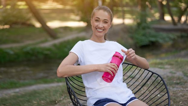 Woman sitting chair drink water red bottle after morning workout. Young athletic female rear view walk asphalt running track park after jogging Healthy lifestyle concept