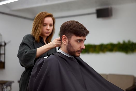Stylish man sitting barber shop Hairstylist Hairdresser Woman cutting his hair Portrait handsome happy young bearded caucasian guy getting trendy haircut Attractive barber girl working serving client