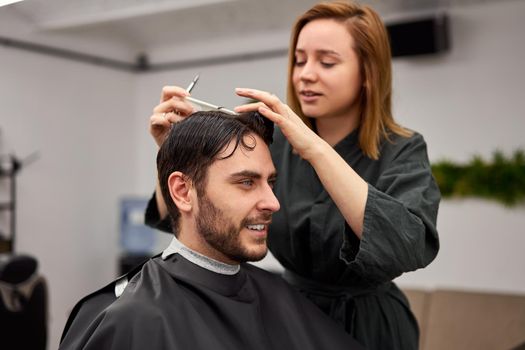 Stylish man sitting barber shop Hairstylist Hairdresser Woman cutting his hair Portrait handsome happy young bearded caucasian guy getting trendy haircut Attractive barber girl working serving client