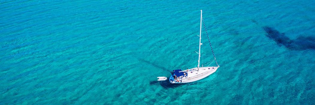 Yacht anchoring in crystal clear turquoise water in front of the tropical island, alternative lifestyle, living on a boat. Aerial view of yacht at anchor on turquoise water, showing luxury, wealth.