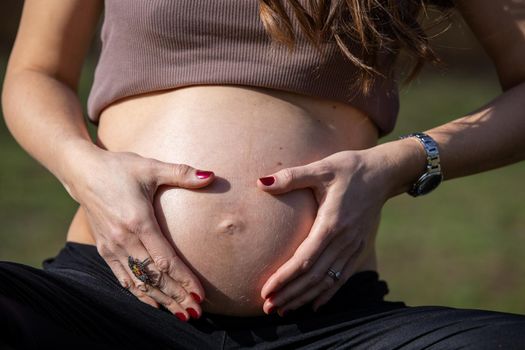 Beautiful pregnant woman feeling happy in nature while taking care of her child. Young expecting mother holding baby in pregnant belly. Maternity prenatal care and woman pregnancy concept.
