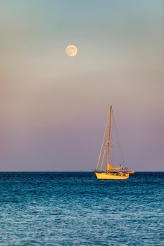 Full moon rising over the water with a small sailing boat in the foreground. Sailing boat with raising moon at sunset. Moon rising over the sea and yacht floating on the water surface. Sardinia, Italy