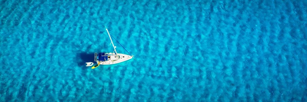 Aerial view of anchored sailing yacht in emerald sea. Aerial view of a boat. Outdoor water sports, yachting. Aerial view of anchoring yacht in open water. Ocean and sea travel and transportation