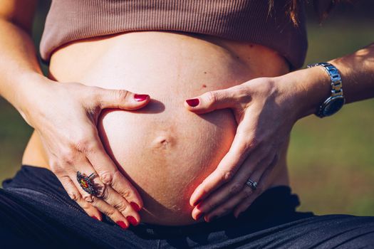 Beautiful pregnant woman feeling happy in nature while taking care of her child. Young expecting mother holding baby in pregnant belly. Maternity prenatal care and woman pregnancy concept.