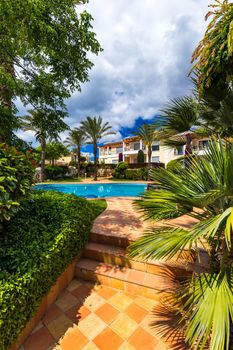 Swimming pool, sun-loungers and palm trees during a warm sunny day, paradise destination for vacations. Backyard swimming pool with garden full of palm trees and flowers. Backyard with swimming pool.