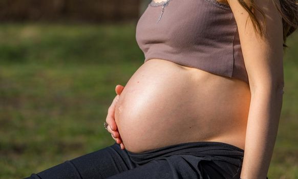 Beautiful pregnant woman feeling happy in nature while taking care of her child. Young expecting mother holding baby in pregnant belly. Maternity prenatal care and woman pregnancy concept.