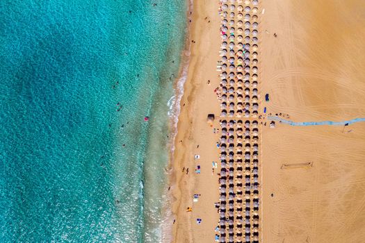 Aerial view of sandy beach with colorful umbrellas, swimming people in sea bay with transparent blue water at sunset in summer. Aerial top view on the beach, umbrellas, sand and sea waves.