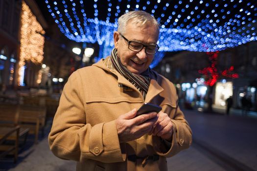 Outdoor portrait of happy senior man in city. He is using smartphone.