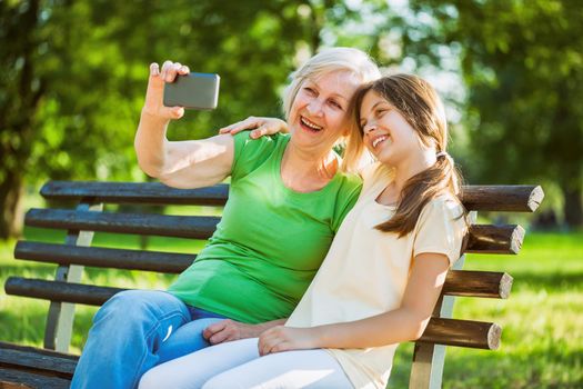 Grandmother and granddaughter are sitting in park and taking selfie.