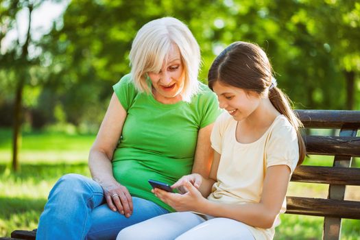 Grandmother and granddaughter are sitting in park and using mobile phone.