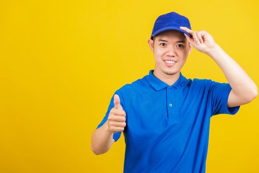 Portrait professional attractive delivery happy man standing he smile wearing blue t-shirt and cap uniform showing thumb up gesture looking to camera, studio shot isolated on yellow background