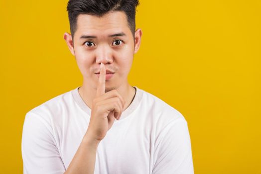 Asian handsome young man holding index finger on lips asking for silence or secret gesturing, studio shot isolated on yellow background, making shh shush gesture concept