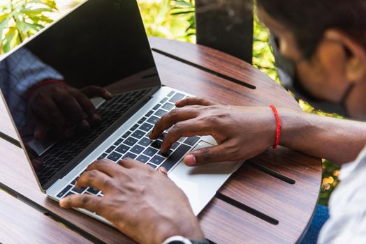 Happy Asian black businessman person or office worker wear face mask protect sitting on desk work from home using laptop computer typing keyboard and connect networking online at the coffee cafe shop