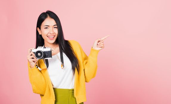 Attractive energetic happy Asian portrait beautiful cute young woman teen excited smiling holding vintage photo camera and pointing finger to side space, studio shot isolated on pink background