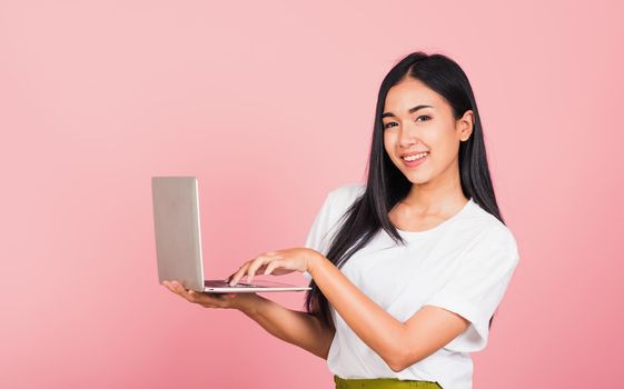 Portrait of happy Asian beautiful young woman confident smiling holding using laptop computer and typing keyboard for online sending email, studio shot isolated on pink background, with copy space
