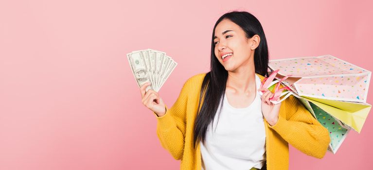 Portrait Asian happy beautiful young woman shopper smile excited holding online shopping bags colorful multicolor and dollar money banknotes on hand in summer, studio shot isolated on pink background