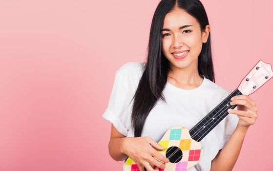 Portrait of happy Asian beautiful young woman teen confident smiling face hold acoustic Ukulele guitar, female playing Hawaiian small guitar, studio shot isolated on pink background, with copy space