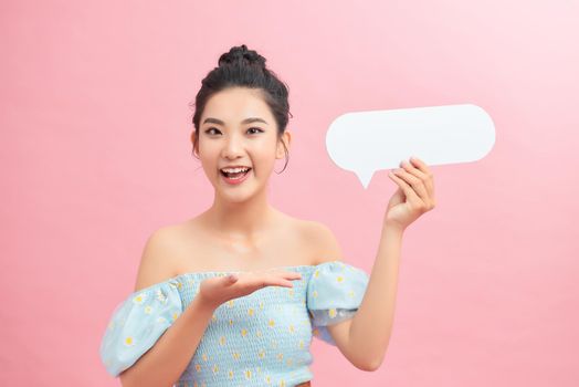 Surprised happy woman holding blank speech bubble and looking at the camera over pink background