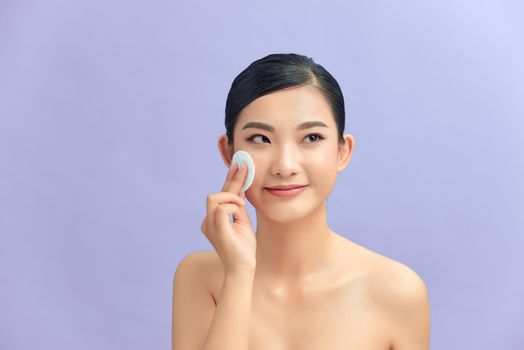 Crop attractive young female cleansing face with a cotton pad on a purple background in studio