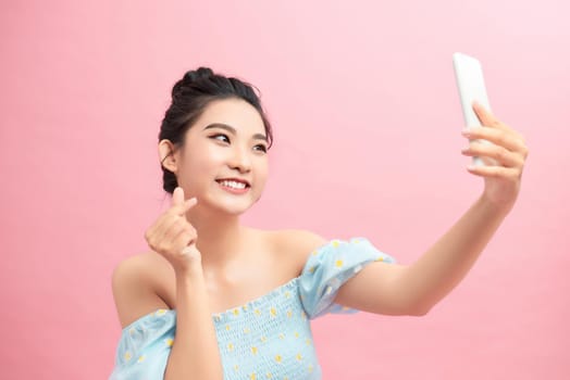 Image of stylish girl showing heart sign and mobile phone screen, like something online, standing over pink background