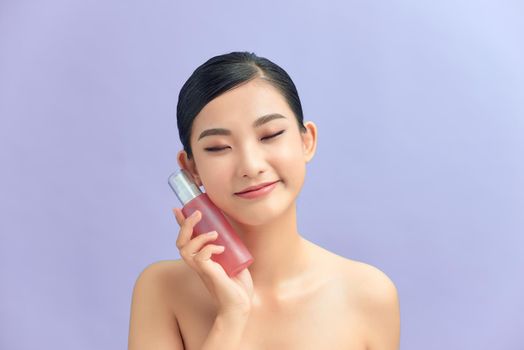 Beauty portrait of a pretty young topless woman wearing makeup isolated over purple background, showing cosmetic skin oil in a bottle
