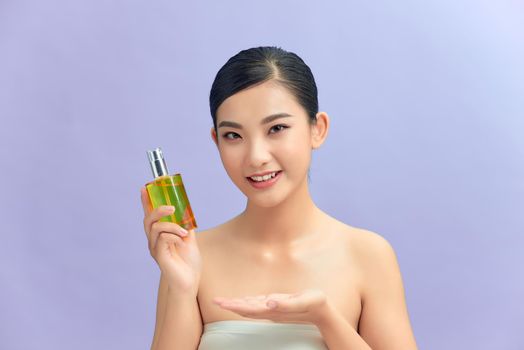 Beauty portrait of an attractive happy healthy topless brunette woman isolated over purple background, showing cosmetic bottle