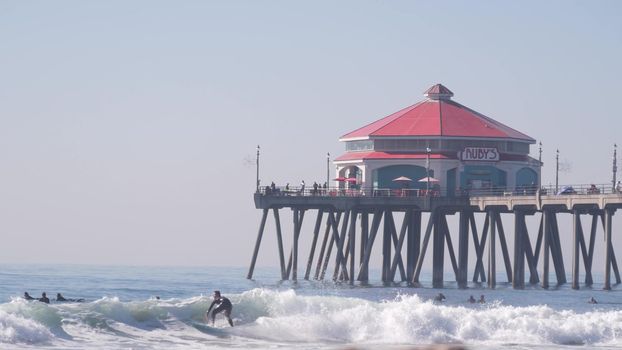 Huntington Beach, California USA - 2 Dec 2020: People surfing in ocean waves, pier on coast. Rubys diner on beachfront boardwalk. Surfers in water, vacations on beach near Los Angeles, Orange county.