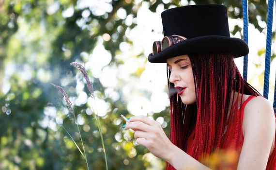 Glamorous girl with scarlet dreadlocks, red swimsuit, black hat and welding glasses posing outdoor with a cigarette 