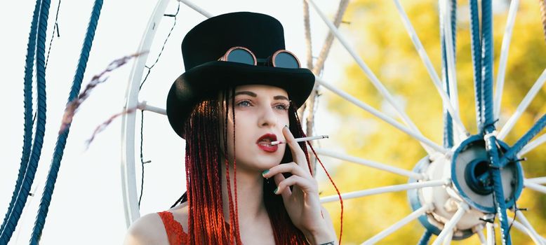 Glamorous girl with scarlet dreadlocks, red swimsuit, black hat and welding glasses posing outdoor with a cigarette 