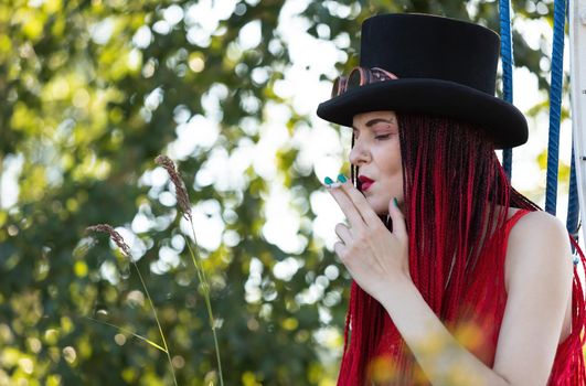 Glamorous girl with scarlet dreadlocks, red swimsuit, black hat and welding glasses posing outdoor with a cigarette 