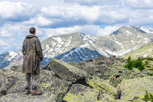 A man stands on a rock among stones against a background of mountains and looks all alone towards the mountain peaks. The scale of man and majestic mountains. View of a man from the back to mountain peaks