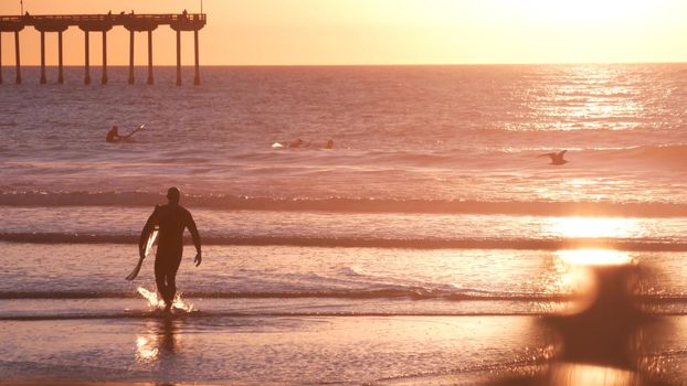 San Diego, California USA - 28 Nov 2020: People surfing by Ocean Beach pier on piles at sunset. Surfers on surfboards in water waves. Vacations on pacific coast or shore. Recreational sport hobby.